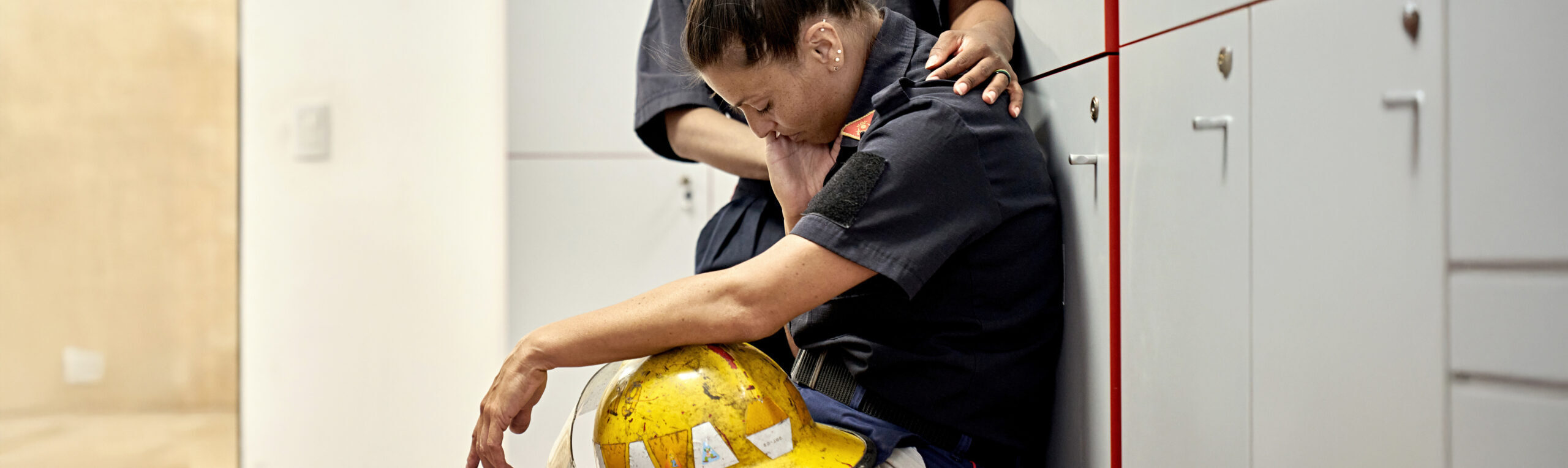 Firefighter in uniform sitting on bench in fire station with helmet in lap, head down, and eyes closed.